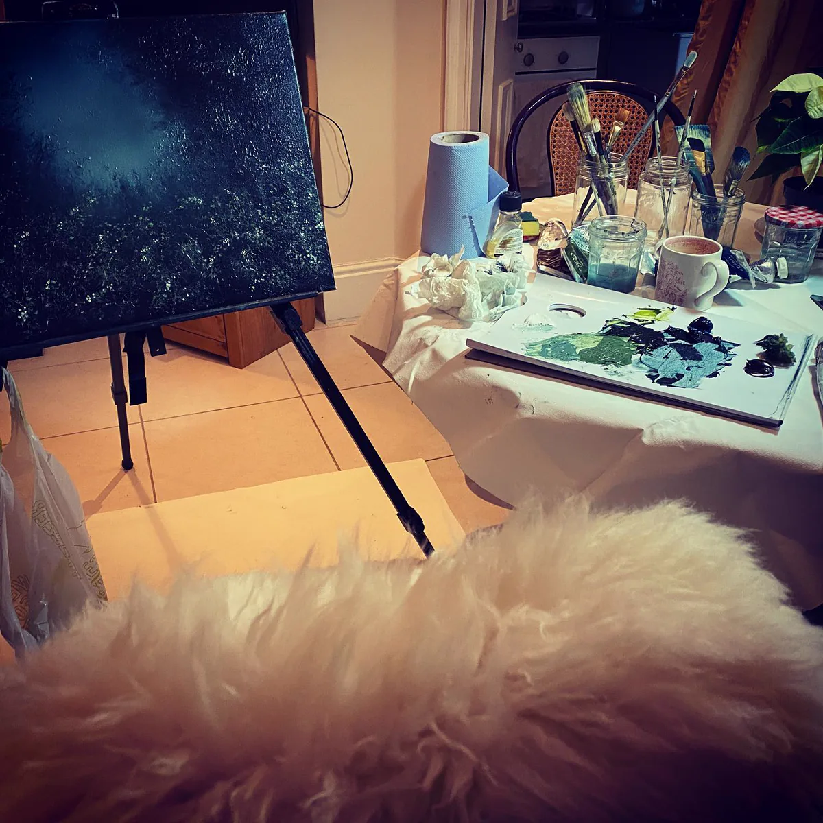 A painting in progress on the easel at home, with brushes and paints on the table